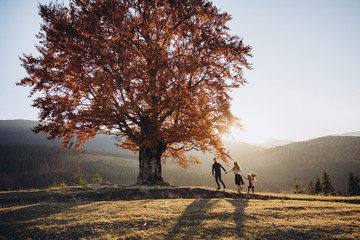 Stylish young family in the autumn mountains. A guy and a girl with their daughter walk together under a large old tree against the background of a forest and mountain peaks at sunset.