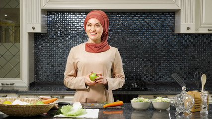 Wall Mural - cheerful woman in hijab blogs culinary show telling subscribers fruit salad recipe standing in home kitchen closeup