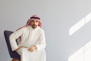 Wall Mural - Pensive arab businessman sitting in a chair thinks looks up on a gray background. Portrait of an attractive arab man.