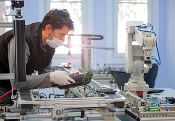 Man with surgical mask on face against COVID-19 ( Coronavirusis ) programming robot arm with control panel which is integrated on smart factory production line. industry 4.0 automation line.