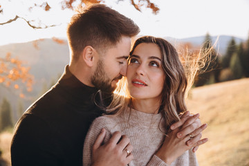 Stylish young couple in the autumn mountains. A guy and a girl hug together under a large old tree on a background of a forest and mountain peaks at sunset.