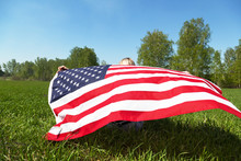 A Close-up Of The American Flag Waving In The Wind Behind The Back Of A Girl Sitting On The Grass In The Forest.