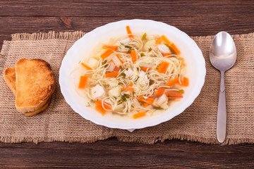 Fresh vegetable soup with pieces of chicken breast and noodles in a white plate served with two slices of toast, angle view