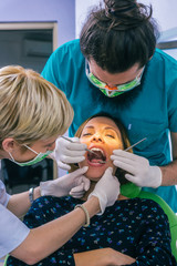 Wall Mural - Close up portrait of a beautiful young lady sitting in the dental chair while stomatologist ( dentist ) with hands in sterile gloves is fixing her teeth.