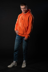 Poster - Young handsome man in jeans and orange hoodie posing while standing on a black background.