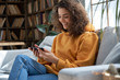 © insta_photos - Happy young hispanic latin teen girl sit on sofa at home holding phone looking at screen watching social media video content, movie or stream, video calling online in mobile app using smart phone.