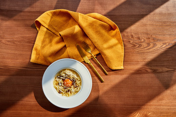 Wall Mural - top view of delicious pasta carbonara served with golden cutlery and yellow napkin on wooden table in sunlight