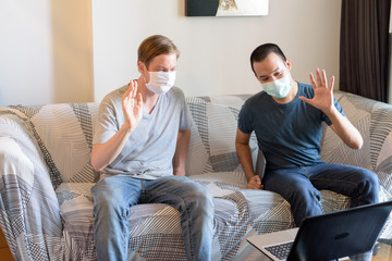 Wall Mural - Two happy multi ethnic men with mask as friends video calling in quarantine at home