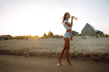 Pretty girl drinking beer while having a party at the beach at sunset. Young woman enjoying on beach holiday. Summer, relax and lifestyle concept.