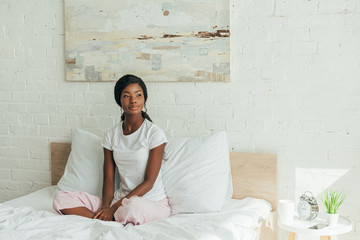 pensive african american girl in pajamas sitting in bed with crossed legs and looking away