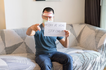 Wall Mural - Young Asian man with mask and face shield showing Stay At Home sign and pointing at camera under quarantine