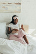 © LIGHTFIELD STUDIOS - african american girl with sleep mask on forehead sitting in bed, reading book and holding cup of coffee