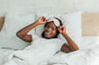 © LIGHTFIELD STUDIOS - smiling african american girl touching sleep mask while lying in bed and looking at camera