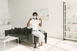© LIGHTFIELD STUDIOS - young african american girl in medical mask showing thumbs up while sitting on grey sofa in kitchen