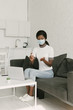 © LIGHTFIELD STUDIOS - young african american freelancer in medical mask spraying antiseptic on hands while sitting near laptop in kitchen
