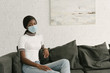 © LIGHTFIELD STUDIOS - pensive african american girl in medical mask sitting on grey sofa at home