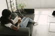 © LIGHTFIELD STUDIOS - High angle view of African American freelancer resting on sofa while reading book near table with laptop