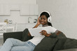 © LIGHTFIELD STUDIOS - happy african american girl in wireless headphones sitting on sofa in kitchen and holding digital tablet