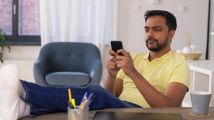 Canvas Print - technology, remote job and people concept - indian man with smartphone resting feet on table at home office