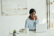 © LIGHTFIELD STUDIOS - attentive african american freelancer talking on smartphone near laptop, wireless headphones and coffee cup