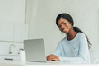 © LIGHTFIELD STUDIOS - cheerful african american freelancer smiling at camera while working on laptop in kitchen