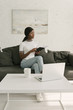 © LIGHTFIELD STUDIOS - thoughtful african american freelancer holding notebook while sitting on sofa near table with laptop