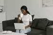 © LIGHTFIELD STUDIOS - young african american blogger sitting on sofa, using laptop and drinking coffee