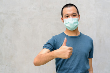 Wall Mural - Young Asian man giving thumbs up with mask for protection from corona virus outbreak outdoors