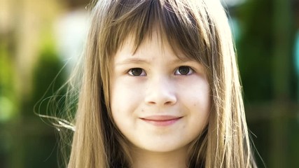 Wall Mural - Portrait of pretty child girl with gray eyes and long fair hair smiling outdoors on blurred green bright background. Cute female kid on warm summer day outside.