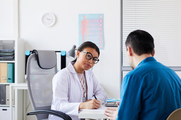 Wall Mural - Young female doctor sitting at the table making notes and listening to patient during his visit at office