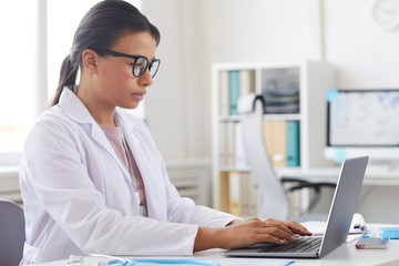 Wall Mural - Female doctor in eyeglasses and in white coat sitting at the table and concentrating on her work on laptop computer