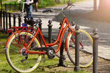 A Bright Red Bicycle Stands At The Fence In A Green Park On A Sunny Day. Moving By Bike Every Day. Bicycle At Street Parking Outdoors. Use Of Eco-friendly And Sports Transport In The City To Move