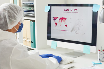 Wall Mural - Female doctor in protective clothing and in protective mask sitting at her workplace and examining the statistics in the world on computer