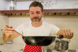 © TheVisualsYouNeed - handsome and happy man in red apron holding cooking pan and wooden spoon smiling cheerful preparing delicious meal in home cook hobby and healthy nutrition