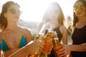  Slender girls cheers and drink beers on the beach at sunset. Young four girl in bikini enjoying on beach holiday.  Summer holidays, vacation, relax and lifestyle concept.