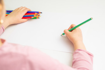 Poster - Kid girl is drawing by green pencil on white paper holding bunch of colored pencils in her hand. Selective focus.