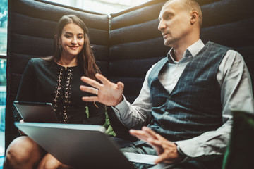 Two business colleagues are talking animatedly while sitting on an office sofa: a smiling businesswoman is listening to her colleague with a laptop that is actively gesturing in a defocused foreground