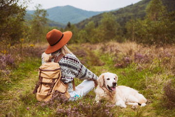 Young hippie blond hair woman in wool sweater with textile backpack and smiling happy golden retriever dog sitting on grass in summer mountains valley. Pets hiking and weekend activities concept.