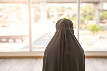 Wall Mural - silhouette of asian muslim Islamic woman praying to allah, back view wearing long black hijab robe, in a private prayer room with light windows shining through, in urban district area and bridges