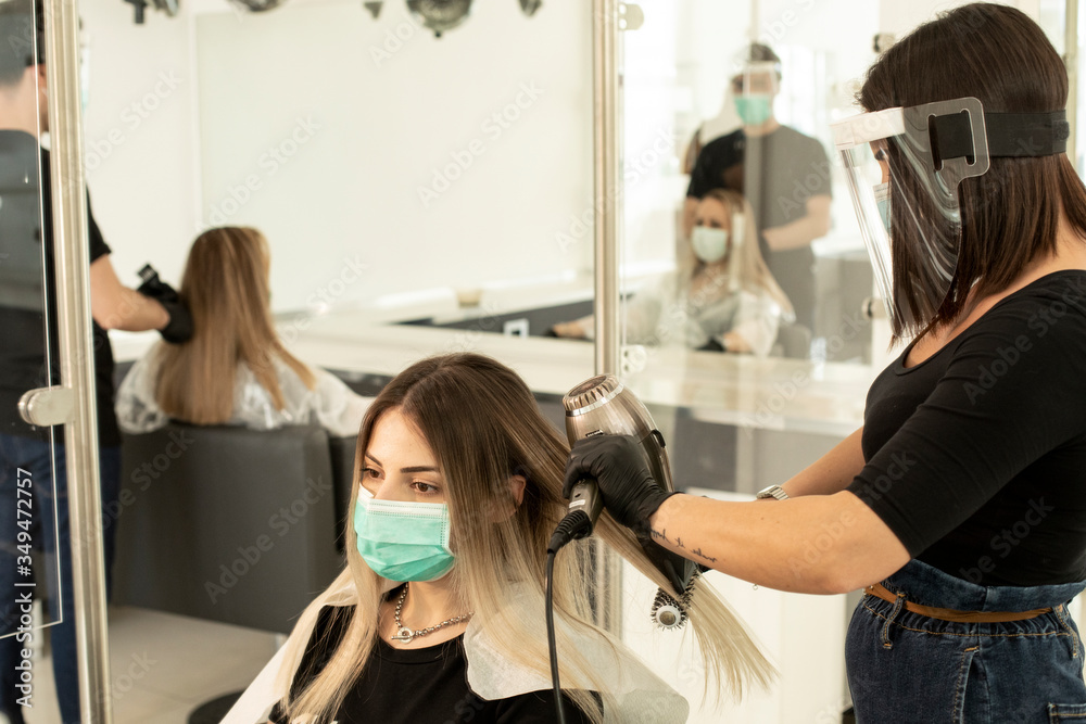 parrucchiera si accinge ad asciugare con un phon i capelli di una di una cliente all'interno di un salone. Entrambi sono equipaggiati di protezioni per evitare contatti e contaminazioni reciproche - obrazy, fototapety, plakaty 