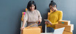 © sutthinon602 - Women packing parcels with sticky tape, Entrepreneur owner using smartphone or laptop taking receive and checking online purchase shopping order to preparing pack product box. Selling online
