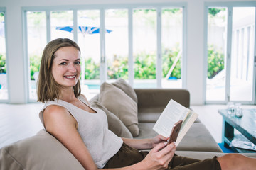 Cheerful young woman on couch with book at home