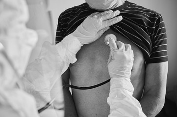 Cropped close-up snapshot of doctor in PPE protective suit examining elderly male patient at home using phonendoscope, checking chest, black and white, COVID-19 concept