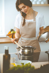 Wall Mural - Young brunette woman cooking soup in kitchen. Housewife holding wooden spoon in her hand. Food and health concept