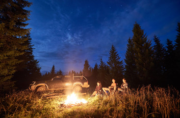 Group of friends near bonfire, pickup offroad truck at night camp in the mountains surrounded by spruce trees under magical starry sky. Car travel and night camping concept.