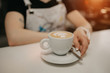 © Roman Tyukin - A female barista holds out a cup of fresh latte with a saucer and spoon to a client in a cafe. A barista preparing a customer order.