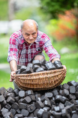 Portrait of an old man picking up a basket full of coal briquettes from a pile in the backyard