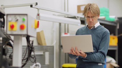 Tracking medium shot of concentrated middle aged engineer in eyeglasses working on laptop computer standing by cnc machine in factory laboratory