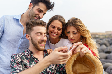 Wall Mural - Group of twentieth young friends are watching a movie on the smartphone together. Smiling happy boys and girls holding a cell phone using social media application outdoor in vacation at the coastline.