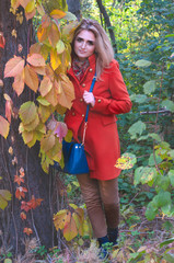 beautiful smile woman in orange coat with bag looking at camera and standing near tree with yellow and green foliage in forest